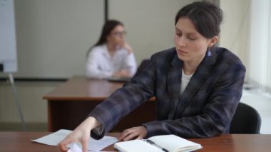Ön planda, bir kadın kağıt belgelere bakar ve kapalı bir masada otururken bir not defterine kalemle yazar. Arka planda, hafif bir defoktus ile bir kız dizüstü bilgisayarda oturuyor.