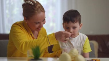 Yakın plan. Mutlu bir anne, oğlunun oturma odasındaki yemek masasında onunla otururken kahvaltı gevreği ve yoğurtla yoğurt besler. Çocuk yemek yiyor.
