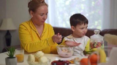 Bir anne kucuk oglunun agzina bir yemek kaşıgi dayadi, onunla oturma odasindaki yemek masasinda otururken ona yoğurtla kahvaltı gevregi yedirmeye çalisiyordu. Çocuk yemeyi reddediyor.