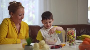 Küçük bir çocuk cam kavanozdan halka şeklinde çok renkli mısır gevreğini alıyor ve yemek masasındaki cam yoğurda ekliyor. Anne, küçük oğlu gibi gülümseyerek izliyor.