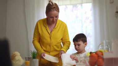 Bir anne küçük oğluna, halka şeklinde renkli tahıl gevreği ile dağınık yemek masasını temizlemesi için kağıt peçete verir. Küçük çocuk masadan tabağa mısır gevreğini süpürüyor.