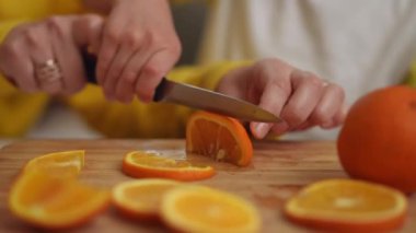 Yakın plan. Bir kadının elleri ve bir çocuğun elleri bir mutfak bıçağıyla portakalı dilimlerken tahta bir tahta kesme tahtasının üzerinde, masada otururken.