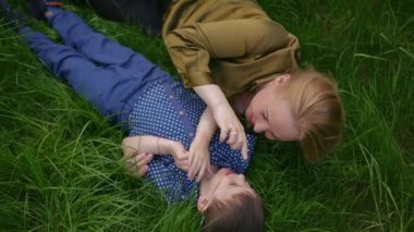 Ağır çekim. Üst Manzara. Mutlu anne ve küçük oğlu, yeşil çimlerin üzerinde sırtüstü yatıyorlar ve bir bahar günü gülümsüyorlar.