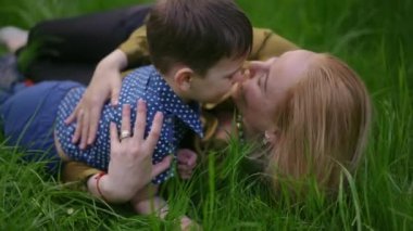 Ağır çekim. Sevecen anne, bir bahar günü yeşil çimenlerin üzerinde uzanırken küçük oğlunu şefkatle kucaklıyor ve öpüyor.