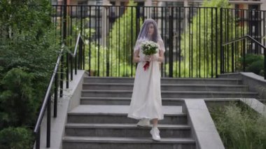 Yüzünde duvak olan gelinlik giymiş genç bir kadın yaz günü yavaşça şehir merdiveninin granit basamaklarından aşağı inerek kameraya bakıyor. Gelin elinde bir buket beyaz papatya tutuyor.