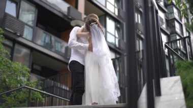Tanınmayan gelin ve damat kucaklaşır ve öpüşür, bir yaz günü merdivenlerin önünde modern bir apartmanın arka planına karşı dururlar.