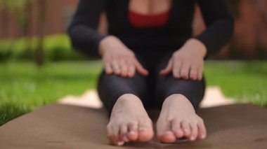 Yakın plan. Eşofman giymiş tanınmayan olgun bir kadın ellerini ayak parmaklarına kadar uzatarak yoga minderinde otururken Asana 'yı canlandırıyor. Güneşli bir bahar gününde dışarıda yoga yapan bir kadın.