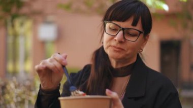 Yakın plan. Gözlük takan bir kadın salata yemekten, plastik çatal ve karton bir tabak tutmaktan, öğle yemeği arasında arka planda bir binayla dışarıda oturmaktan hoşlanır.