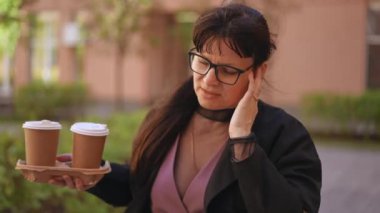 Ağır çekim. Gözlük takan bir kadın bir apartmanın avlusunda yürüyor ve elinde iki plastik bardakla elinde bir karton tutucu taşıyan kulaklıkla konuşuyor.