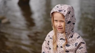 Renkli yağmurluk giymiş bir çocuk sakin suyun kenarında duruyor ve doğadaki mükemmel ve huzurlu anı yakalıyor.