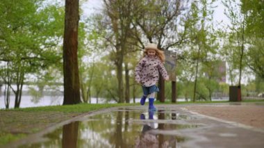Neşeli, kaygısız bir çocuk, yemyeşil bir parkta yağmurlu güzel bir günün tadını çıkararak, su birikintisinde mutlulukla sıçrıyor.