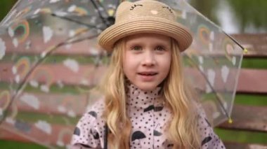 Parlak sarı yağmurluk ve hasır şapka giyen neşeli ve neşeli bir çocuk berrak bir şemsiyeyle mutlu bir şekilde poz veriyor.