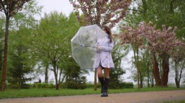 Genç bir kadın elinde renkli bir şemsiyeyle bahar parkında yürür çiçek açan kiraz ağaçlarının altında.