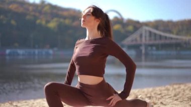 Nehir kenarındaki kumlu bir sahilde yoga yapan bir kadının sakin bir sahnesi. Etrafı canlı ve gür doğa ile çevrili.