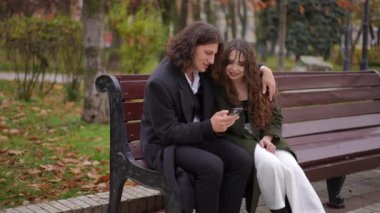 Bir çift parktaki bankta mutlu bir şekilde akıllı telefon kullanırken samimi ve sevgi dolu bir anın tadını çıkarıyor.