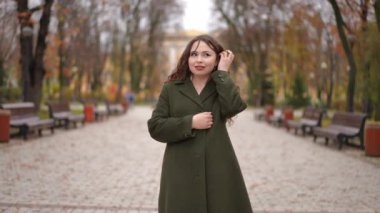 Çevresi güzel bir sonbahar parkı manzarasıyla çevriliyken bir kadın canlı yeşil bir ceketle güvenle ayakta duruyor.