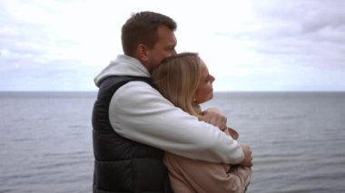 Ağır çekim. Sevgi dolu bir çift, sakin, sakin bir denizin kenarında, yumuşak, bulutlu bir gökyüzünün altında kucaklaşarak güzel ve hassas bir anı paylaşır.