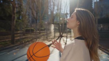 Genç bir kadın, canlı şehir manzarası ve parlak güneş ışığıyla çevrili bir basketbol topuyla güvenle dışarıda duruyor.