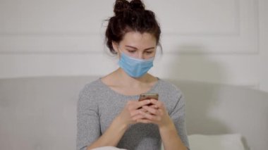 Mavi bir maske takan genç bir kadın evde otururken akıllı telefonuyla etkileşime giriyor.