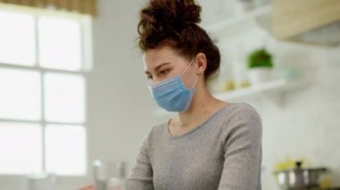 Maske takan genç bir kadın, parlak ve davetkar bir mutfakta çalışırken derin ve düşünceli bir şekilde yansıtır.