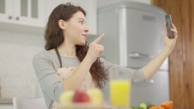 Neşeli ve hayat dolu genç bir kadın, parlak tasarlanmış, şık mutfağında eğlenceli bir selfie çekiyor.