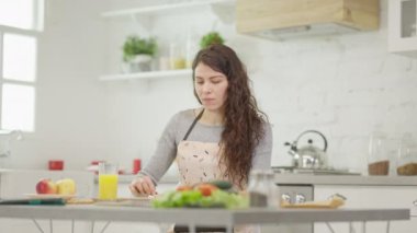 Önlük takan odaklanmış genç bir kadın şık, modern bir mutfakta sağlıklı bir yemek için taze malzemeler hazırlıyor.