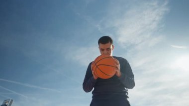 Genç bir adam açık havada güzel mavi bir gökyüzünün altında basket oynamaya hazırlanıyor.