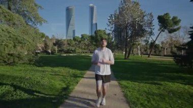 Bir koşucu, arka planında modern gökdelenler olan güzel, yemyeşil bir şehir parkında mutlu bir şekilde güneşli bir günün tadını çıkarıyor.