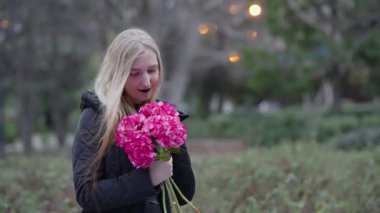 Huzurlu bir park ortamında, genç bir kız neşe saçan bir buket pembe çiçek tutar.