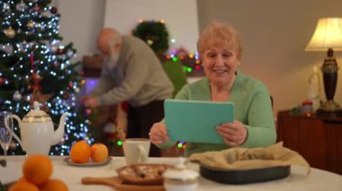 Yaşlı bir kadın festival masasında tablet kullanırken gülümsüyor, sıcak bir bayram anının tadını çıkarıyor. Arkasında, bir adam güzel aydınlatılmış bir Noel ağacının yanında süslemeler düzenliyor..