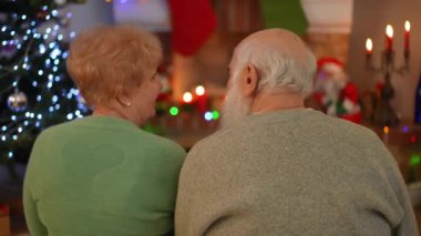 Yaşlı bir çift, aydınlık bir Noel ağacının yanında birlikte otururken sakin ve samimi bir anı paylaşıyor. Şenlikli atmosfer ve ılık dekorasyonlar bir sevgi ve birliktelik hissi yaratıyor..