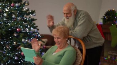 Yaşlı bir çift, süslü bir Noel ağacının yanında oturmakta olan bir video görüşmesi sırasında gülümseyip el sallıyor. Neşeli ve sıcak tatil atmosferi aile bağının neşesini yansıtıyor..