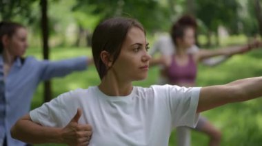 Beyaz kıyafetli bir yoga öğrencisi açık havada, yemyeşil bir parkta Warrior II pozu veriyor. Huzurlu ortam, grup yoga yaparken odak ve farkındalığı arttırır.