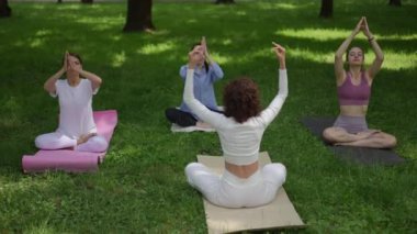 Bir grup kadın Lotus Poz 'unda oturur, sakin bir park ortamında meditasyon ve farkındalık üzerine odaklanırlar. Bu yoga seansı dengeyi, rahatlamayı ve doğayla bağlantıyı vurguluyor.