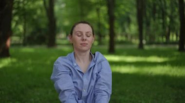 Mavi gömlek giyen genç bir kadın yeşil bir parkta enerjik yoga hareketleri sergiliyor. Elleri havada ve gözleri kapalı, odaklanmayı ve kararlılığı yönlendiriyor. Özgüvenli ifadesi ve hareketli