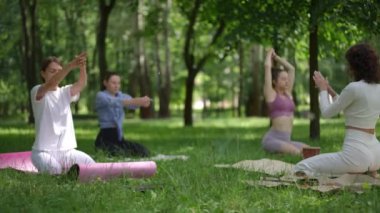 Bir grup kadın yemyeşil bir parkta canlı bir oturum sırasında enerjik bir şekilde yoga egzersizleri yapıyor. Beyazlı öğretmen uygulamayı yönetirken, katılımcılar esneyip odaklanıyorlar. Onların...