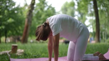 Beyaz giyinmiş genç bir kadın, pembe bir minderde yoga yaparken Cat Poose ya da Marjaryasana 'yı gösteriyor. Omurgası yukarı doğru eğiliyor ve nefes kontrolü ve omurga hizasına odaklanıyor. Poz.