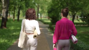 Gölgeli bir park yolunda yürüyen ve formda figürlerini sergileyen iki kadının arka görüntüsü. Biri bej bir yoga minderi taşırken, diğeri ise katıksız pembe bir bluz giyer ve tayt giyer.