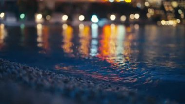 Sesli izleyin. İspanyol sahilinin sakin sularında parıldayan altın ışıkların yumuşak Bokeh yansımalarını yakalayan sakin bir akşam sahnesi. Nazik dalgalar çakıl taşlı kıyı şeridiyle birleşir