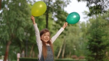 Neşeli ve neşeli bir kız, canlı bir park ortamında bir demet yeşil ve sarı balon tutuyor.