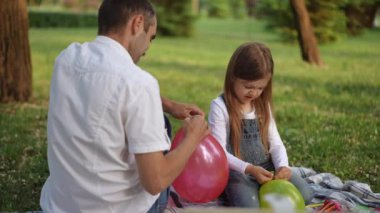 Huzurlu bir parkta renkli balonlar hazırlayan bir baba ve kız arasında paylaşılan yürek ısıtan bir an.