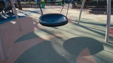 Tek bir siyah salıncak, sağlam zincirlerle asılı duran boş bir oyun bahçesinde asılı duruyor. Güneş ışığı, ağaçlar ve oyun ekipmanlarıyla çevrili renkli lastik yüzeyde çarpıcı gölgeler yaratır. Şey...