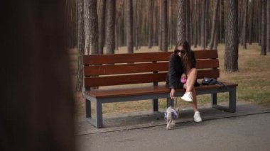 Siyah kıyafetli şık bir kadın orman parkındaki ahşap bir bankta oturuyor ve Yorkshire Teriyeri 'ni kucağında tutuyor. Güneş gözlüğü takarak şık çantasıyla huzurlu bir ortamın tadını çıkarıyor.