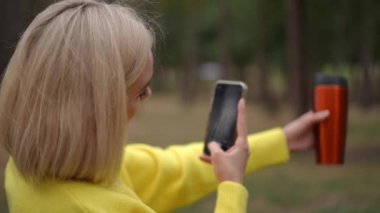 Açık havada fotoğraf çekmek için elinde akıllı bir telefonla gülümseyen sarı bir kazak giyen bir kadının yakın çekimi. Diğer elinde parlak turuncu bir termos tutuyor, rahat ve neşeli bir hava katıyor.