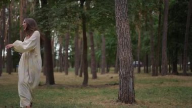 Hafif bir elbise giymiş neşeli bir kadın sakin bir ormanın ortasında ağır çekimde zarif bir şekilde dans eder. Uzun saçları hareketleriyle sallanıyor ve gülüşü mutluluk saçıyor.