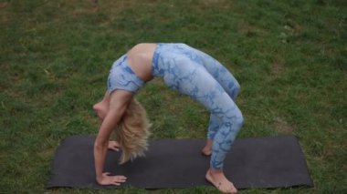 Mavi desenli yoga kıyafetli sarışın bir kadın açık havada siyah bir paspasın üzerinde zarif bir sırt üstü poz veriyor. Sahne onun esnekliğini ve gücünü doğal yeşilliğin ortasında yakalıyor.