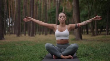 Yoga yapan odaklanmış bir kadın huzurlu bir orman ortamında yoga yapıyor, yoga minderinde bacak bacak üstüne atmış bir pozisyonda oturuyor. Ellerini kaldırır ve dua pozu verir. Sakinlik ve konsantrasyon yayar.