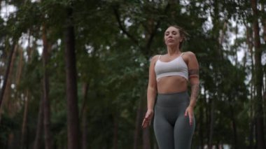 Beyaz spor bluzlu ve gri taytlı bir kadın sakin bir ormanda yukarı doğru uzanıyor. Duruşu güç ve odaklanma gösteriyor. Açık hava ortamında esnekliği ve farkındalığı vurguluyor.