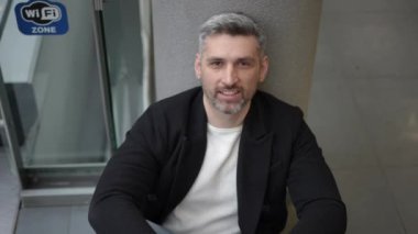Bir alışveriş merkezinin zemininde oturan koyu renk ceketli ve günlük kıyafetli bir adamın yakın plan fotoğrafı. Kamera, Kara Cuma satışlarına katıldıktan sonra gevşemiş ifadesini yakalamak için yakınlaşıyor.