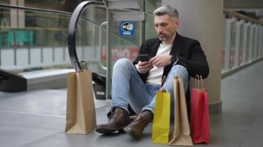 Koyu renk ceketli ve kotlu bir adam alışveriş merkezinin zemininde oturuyor, akıllı telefonunu açık bir Wi-Fi bölgesinde kullanıyor. Etrafı renkli alışveriş torbalarıyla çevrili bir şekilde yürüyen merdivenin yanında mola veriyor.